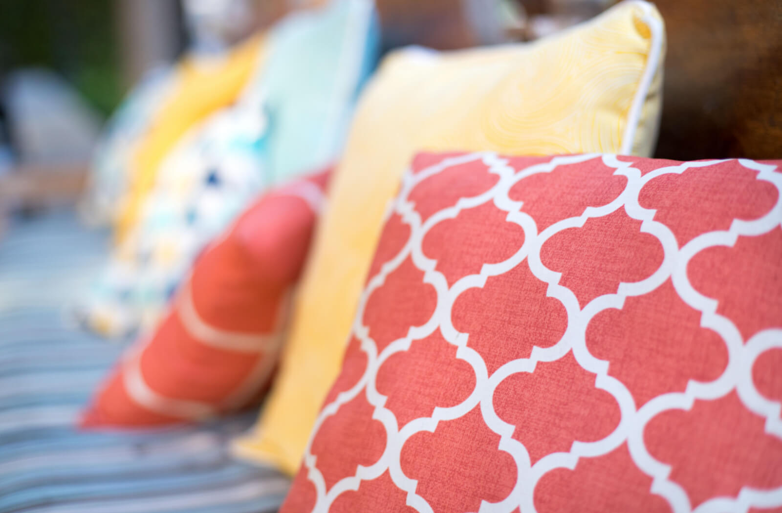 A close up of orange and yellow patterned throw pillows on an outdoor bench with a blue, striped cushion underneath the pillows.