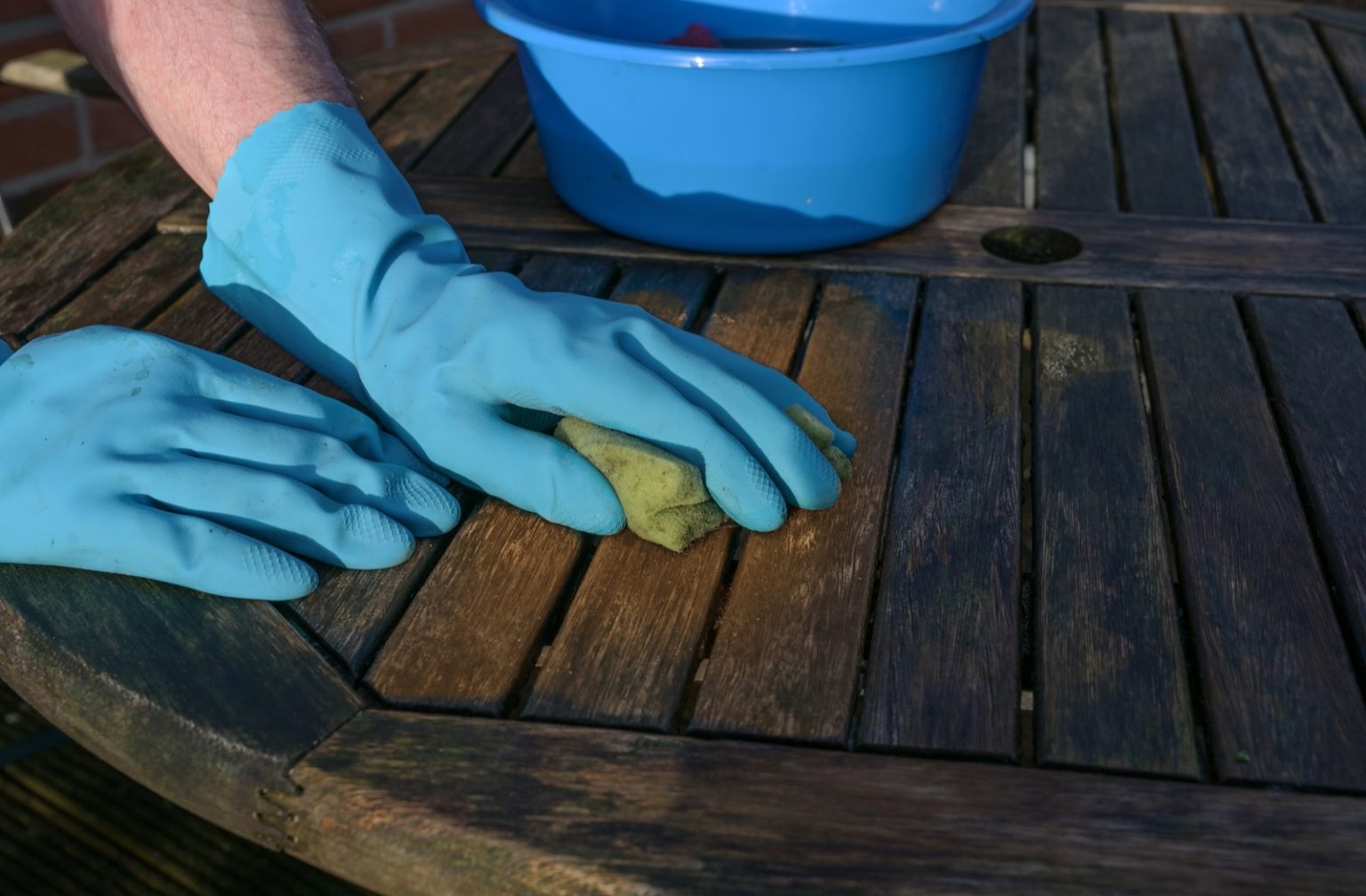 A person wearing blue rubber gloves using a sponge with soapy water to clean their dirty, wooden outdoor patio furniture